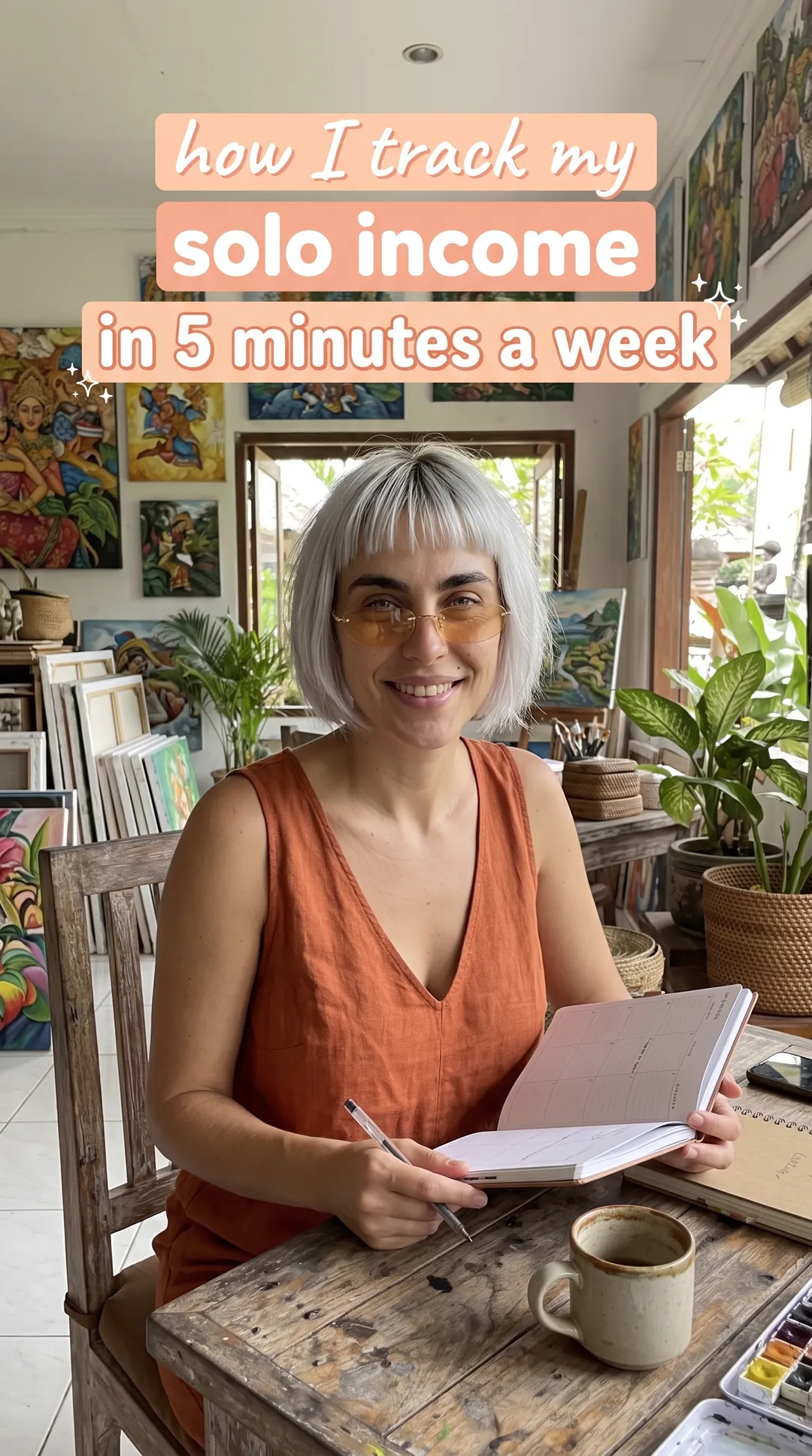 Kaia in a rust sleeveless top writing in a notebook at a wooden table in a colorful studio, Annual Budget Spreadsheet ad
