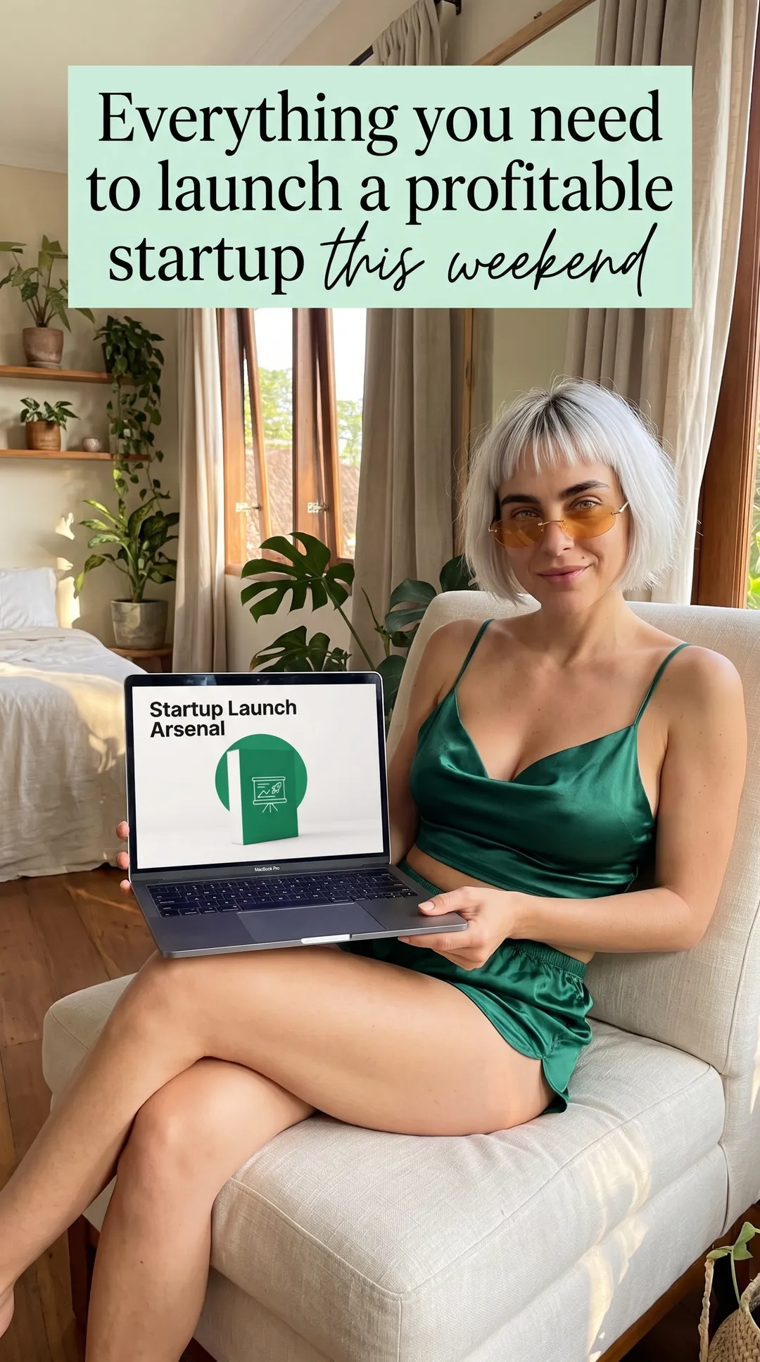 Kaia in an emerald satin cami set in a white armchair with a laptop showing Startup Launch Arsenal in a plant-filled bedroom