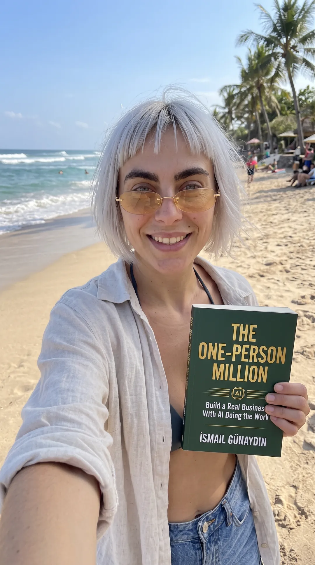 Kaia in a white linen shirt taking a beach selfie with The One-Person Million paperback